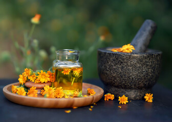 glass bottle essential oil of calendula and fresh calendula flowers leave on a wooden plate and a granite mortar on a black table with a blurred background. Concept: spa, aromatherapy and beauty. - obrazy, fototapety, plakaty