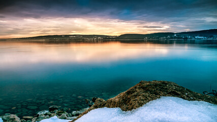 Wunderschöner Sonnenaufgang am Bodensee mit Schnee am Seeufer und Steinen am Wasser 