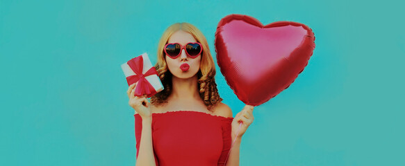 Portrait of lovely woman with red heart shaped balloon and gift box blowing lips on a blue background