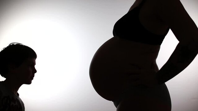 Silhouettes of a pregnant woman and a child kissing mom's belly on a white background. Waiting for the birth of a sibling. Black and white.
