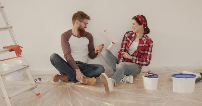 Young happy married couple sitting on the floor on background of apartment during the repair and playfully fighting with paint rollers.