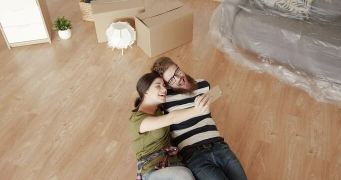 Two happy young people lying on the floor in new apartment and taking selfie on mobile phone making faces.