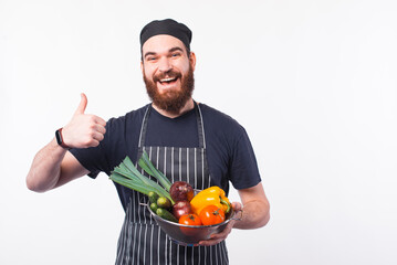 Photo of happy bearded chef man in uniform holding vegetables in bowl.