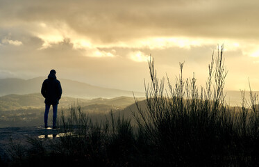 young man in coat, cap and jeans on top of a mountain watching the sunrise views. teenager thinking on top of a mountain in winter at sunset. green and rocky mountains of galicia.