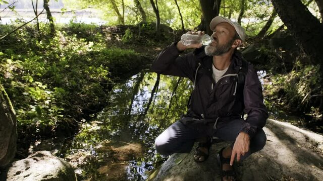 A tired traveler by a stream drinks dirty water from a plastic bottle.