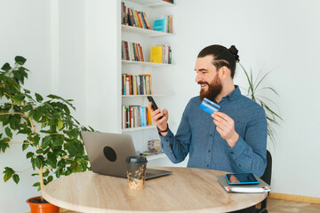 Cheerful man working in office and using credit card and smartphone.
