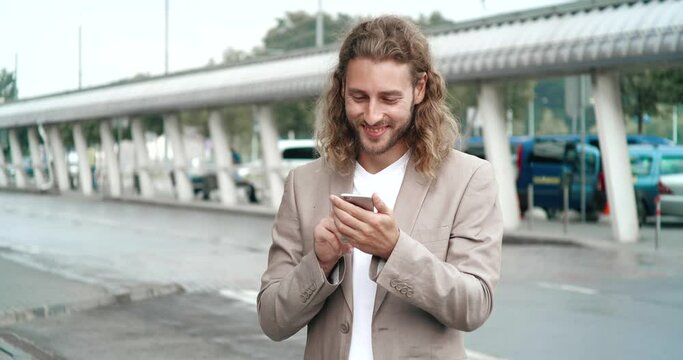Businessman is standing near office center and browsing his Phone. Handsome Young Businessman is having Good Mood, looking satisfied. Businessman is walking in Raing, smiles. People.