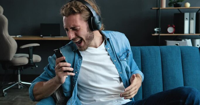 Caucasian Bearded Hipster on the bed and listening to Music in Headphones. Young Man enjoying cool Music on Couch in Loft Living Room. Playfully Mood at Home. FunnyMan. Hobby. Apps.