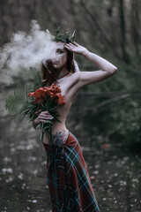 Charming LGBT+ girl witch poses in a gloomy summer forest with tiger lilies in her hands. Young Wiccan woman in a misty haze. Stylish sensual 90's woman. 30 year old beauty in the woods