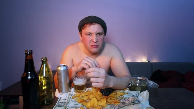 Redneck in a hat and with a naked torso eats chips and scatters money