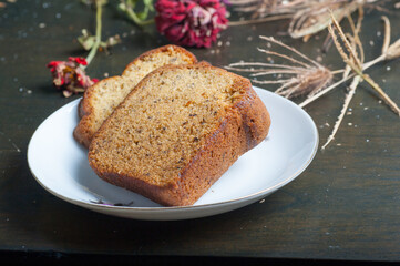 two slices of banana bread on a small plate with a wooden chopping board and garnish with flowers and straw