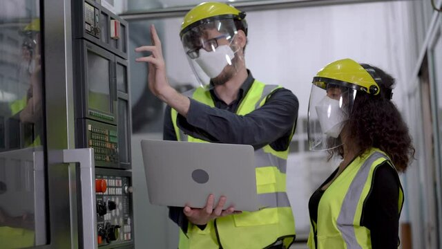 Factory engineer workers with face mask and face shield protection checking and adjusting machine panel with laptop in hi-tech factory. New normal life of people in workplace of industrial business.