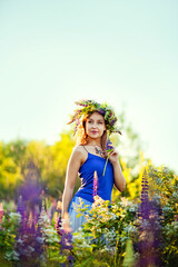 Naklejka na ściany i meble A girl with a flower wreath on her head sits at sunset in a field with purple lupin flowers. Image with selective focus, toning, and noise effect