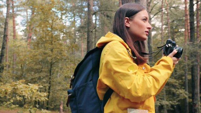 Young beautiful girl tourist in yellow jacket travels alone in woods and takes photos of nature. Caucasian woman is engaged in hobby traveling.