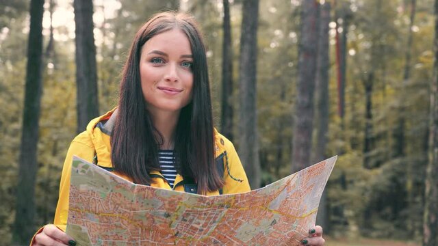 Portrait of happy tourist girl standing on sunny forest glade holding map in her hands looking at camera and smiling.