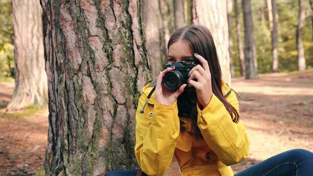 Beautiful girl photographer in yellow jacket is resting in woods under tree and taking photos on camera enjoying nature.