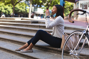 African american woman with lowered face mask drinking coffee while sitting on the stairs outdoors