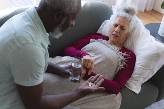 Senior caucasian woman lying sick on couch man passing medicine and water in living room
