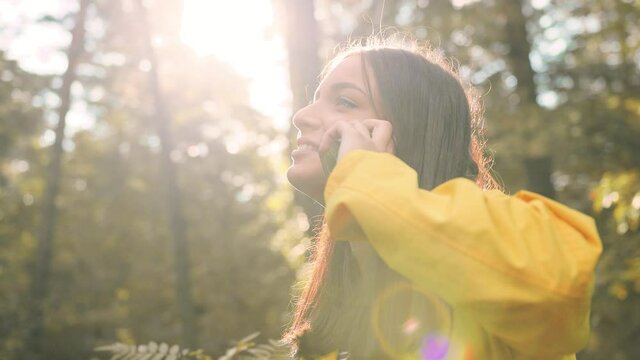 Young girl in yellow jacket stands alone in the middle of forest in sunlight with paper map in her hands and talks on mobile phone.