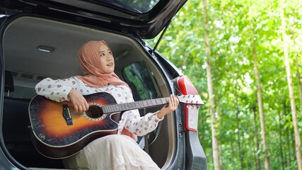 asian woman tavelling wearing hijab playing guitar with a car