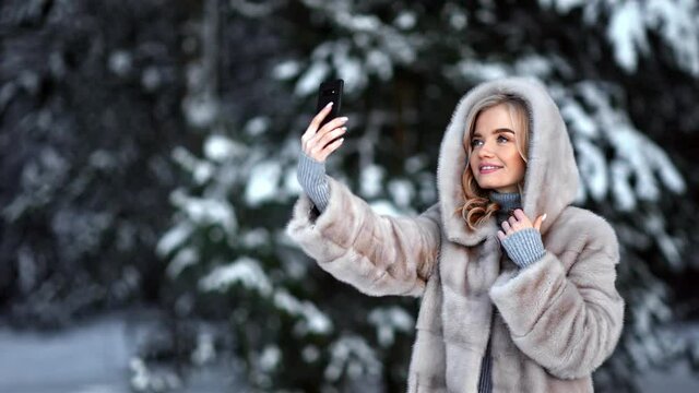 Fashionable lady posing taking selfie in fur coat use mobile at forest. Medium shot on RED camera