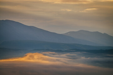 Scenic dawn mountain landscape with golden low clouds in valley among blue mountains silhouettes under cloudy sky. Sunset or sunrise scenery with low clouds in mountain valley in illuminating color.