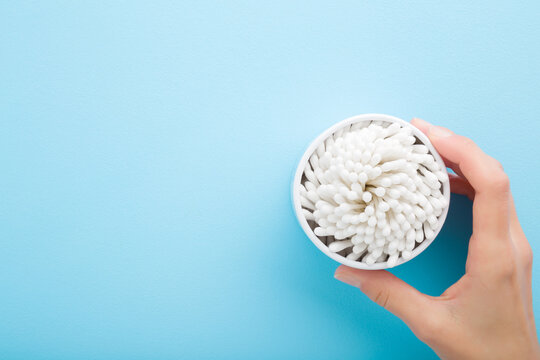 Young Adult Woman Hand Holding Opened Container With Sticks Of White Cotton Bud On Light Blue Table Background. Pastel Color. Closeup. Empty Place For Text. Top Down View.