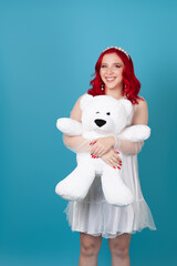 happy cheerful young woman woman with red wavy hair in white dress hugs big white teddy bear isolated on blue background.