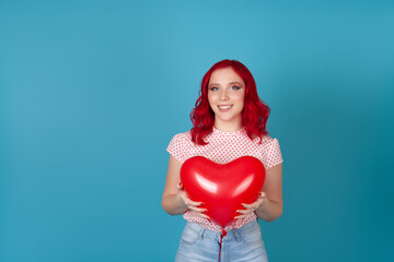 Fototapeta na wymiar close up of a smiling cute young woman with red hair holding a red heart-shaped flying balloon isolated on a blue background with both hands.