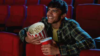 Portrait of cheerful young guy smiling, holding a drink and popcorn basket while watching movie...