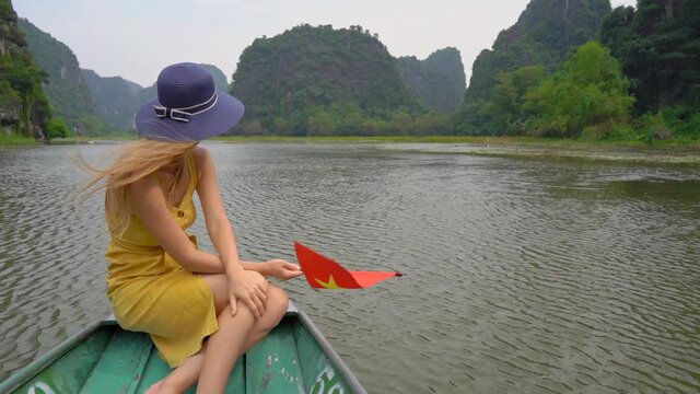 Slowmotion shot of a young woman holding a vietnamese flag in her hand has a river trip among spectacular limestone rocks in Ninh Binh, a tourist destination in northern Vietnam. Travel to Vietnam
