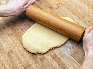 Hands of a caucasian woman is rolling out the dough with a rolling pin on a wooden board.