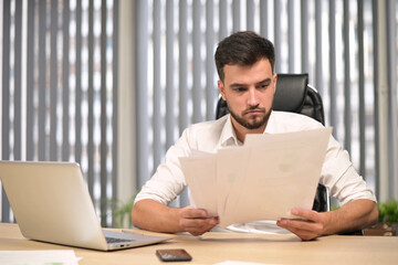 Young head of the company examines the reports printed on paper and is perplexed