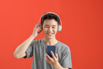 Asian man with headphones listening music using smartphone app while standing over isolated red background