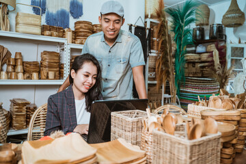a male businessman standing next to a female secretary who sits while working on a laptop at a...