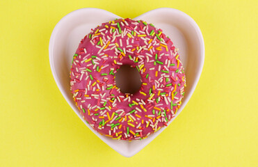 Round sweet pink donut in a heart-shaped plate on a yellow background. Sweet symbol for valentine's...