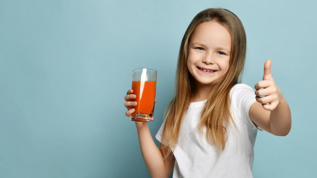 Cheerful happy small cute girl in white t-shirt standing with glass of fresh fruit carrot or orange juice showing thumbs up sign on blue background, copy space. Happy childhood, healthy eating
