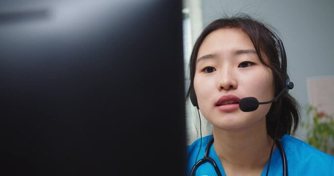 Close up of hard-working Asian woman in blue medical uniform consulting patients online. Young female nurse in headphones conferencing on computer during quarantine in hospital. Medicine.