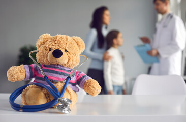 Cute soft teddy bear doctor with stethoscope sitting on table at pediatric clinic or children's medical center. Kids checkup visit to hospital, health maintenance concept. Blurred empty space for text