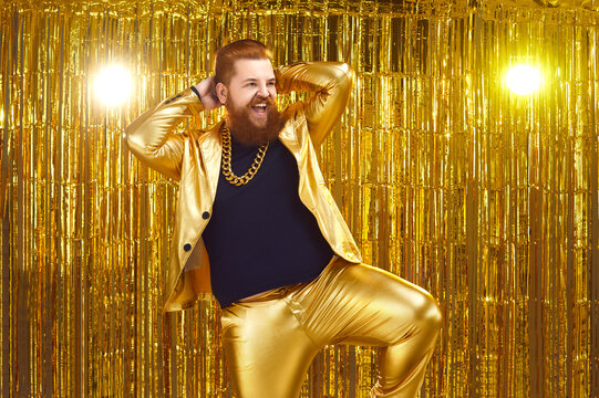 Happy confident young man with ginger beard and mustache wearing funny golden suit with bling gold chain necklace performing on stage at night club disco party, dancing, singing songs and having fun