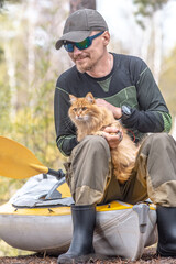 tourist man sits on the shore next to the boat and tent and holding red cat