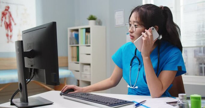Hard-working Asian woman in glasses calling collegue and checking reports on computer. Adult female nurse documenting, typing on keyboard and talking on phone. Gadgets, medicine, technology.