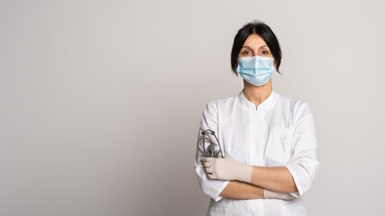 Female doctor or scientist wearing protective facial mask standing with crossed arms over grey background. Place for ad