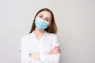 Young woman in medical mask with her arms crossed looks at the camera, white background