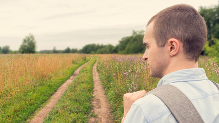 Young traveler with a backpack goes along the road to the field, the forest in the distance, beautiful Russian landscape, rear view, 16:9