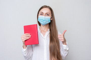 Woman student in a medical mask with a red book on a white background, girl shows a thumbs up