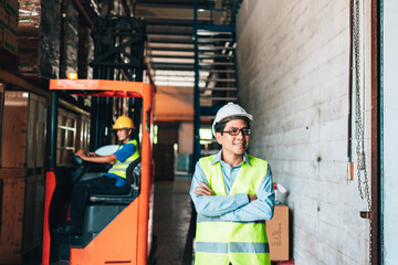 Portrait of asian man. warehouse Smiling worker standing with arms crossed in large warehouse with goods. in background driver at Warehouse forklift loader works with goods.