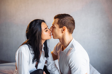 Beautiful passionate couple having sex on the bed. Cheerful romantic lovers hugging. The boyfriend and girlfriend sit cross-legged on a crisp white bed, illuminated by warm yellow light in white room
