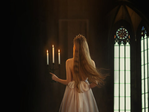 Mysterious art medieval girl princess walks in dark gothic room. Woman queen is holding candlestick with burning candles in hand. Dress with open back long loose blonde hair flying in motion. Go away
