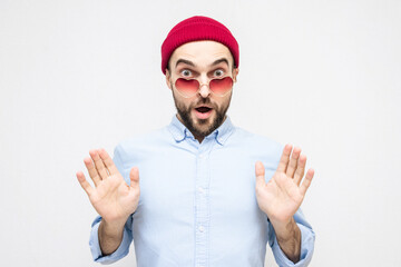 Dazed bearded lover shows stop sign with palms, man wears pink heart-shaped glasses, portrait, white background